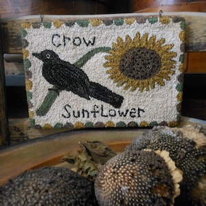 May include: A rectangular, textured wall hanging with a crow perched on a branch next to a sunflower. The words "Crow" and "Sunflower" are stitched on the cream-colored background. The artwork is framed with a scalloped border.