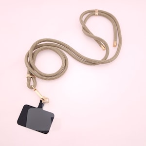 May include: A khaki colored rope phone lanyard with a black phone holder. The lanyard has gold metal clasps.