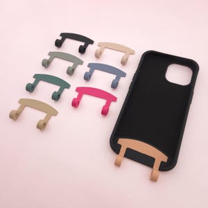 Könnte beinhalten: Ein Satz von acht Handyhüllen-Griffen in verschiedenen Farben, darunter Schwarz, Beige, Grün, Grau, Pink und Blau. Die Griffe sind so konzipiert, dass sie an der Rückseite einer Handyhülle befestigt werden, um einen sicheren Halt zu gewährleisten. Die Griffe sind auf einem rosa Hintergrund abgebildet.