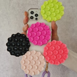 Könnte beinhalten: Eine transparente Handyhülle mit einer Vielzahl von bunten, runden Saugnapf-Handyhaltern. Die Halterungen sind in den Farbtönen Pink, Grün, Schwarz und Pfirsich gehalten. Ein lila Schlüsselband ist an einer der Halterungen befestigt.