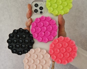 May include: A clear phone case with a variety of colorful, round, suction-cup phone grips attached. The grips are in shades of pink, green, black, and peach. A purple lanyard is attached to one of the grips.