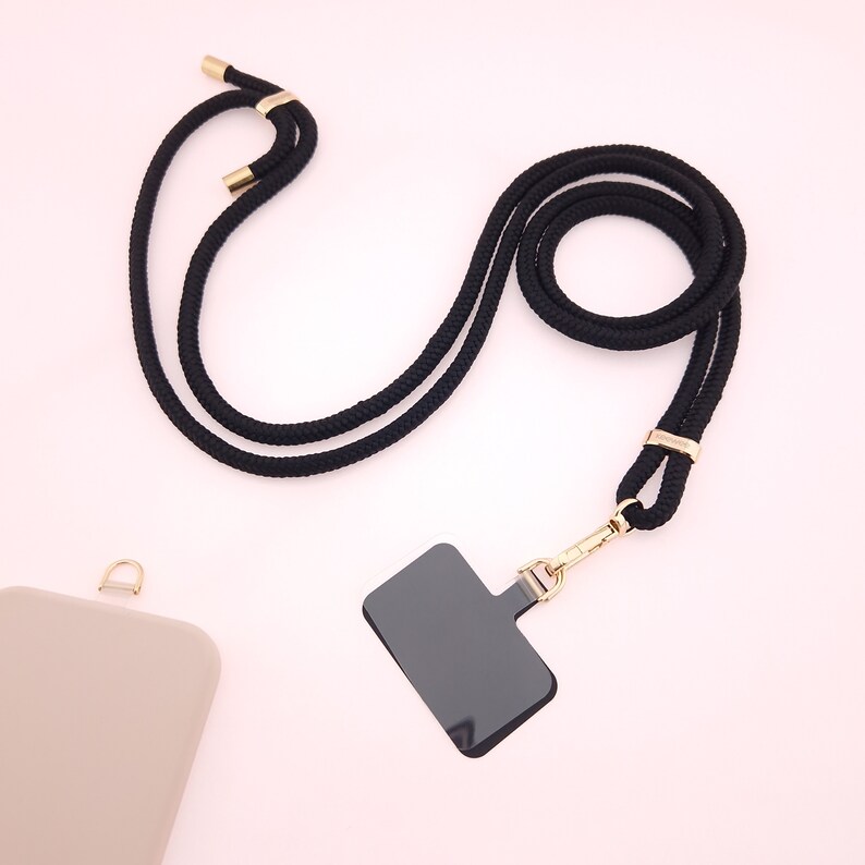 May include: Black phone strap with a gold clasp and a black rectangle phone holder. The strap is made of a thick, braided cord.