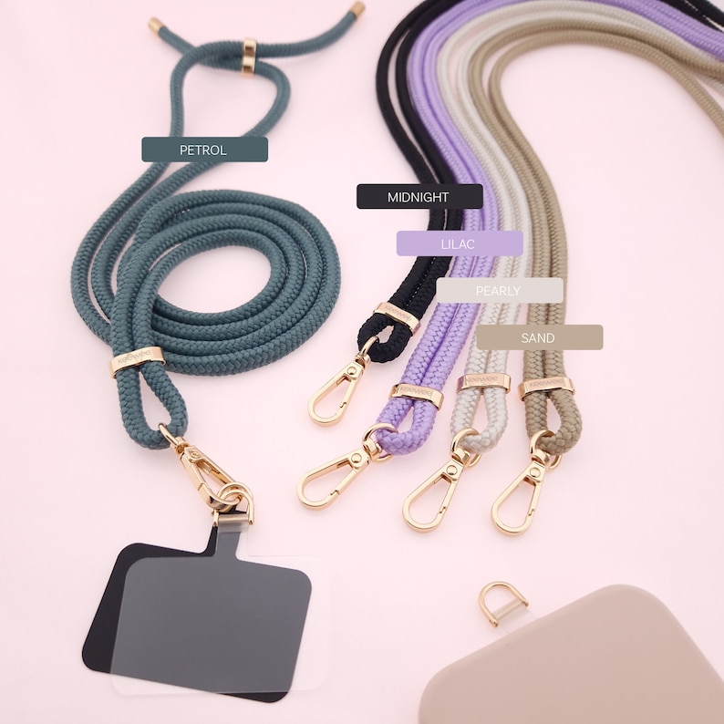 May include: A set of six phone straps in various colors, including petrol, midnight, lilac, pearly, and sand. The straps are made of a braided rope material and have gold-colored clasps. The straps are displayed on a pink background.