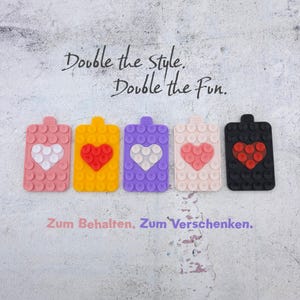 Könnte beinhalten: Fünf farbige Handyhalterungen mit Herzmotiven. Die Halterungen sind rosa, gelb, lila, hellrosa und schwarz. Der Text "Double the style. Double the Fun." steht darüber.