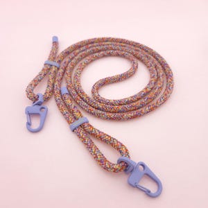 Könnte beinhalten: Ein farbenfrohes geflochtenes Lanyard mit zwei lila Clips. Das Lanyard besteht aus verschiedenen Farben, darunter Rot, Orange, Gelb, Grün, Blau und Lila.