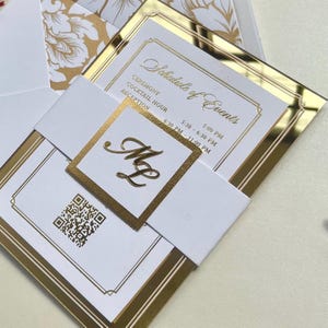 K&ouml;nnte beinhalten: Eine wei&szlig;e Hochzeitseinladung mit goldenen Akzenten und einem spiegelnden Hintergrund. Die Einladung enth&auml;lt den Text "Schedule of Events" und eine Liste von Veranstaltungen mit Uhrzeiten. Die Einladung enth&auml;lt auch ein goldenes Monogramm mit den Initialen "M&M".