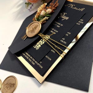 K&ouml;nnte beinhalten: Schwarze und goldene Hochzeitseinladung mit einem Wachssiegel und einem QR-Code. Die Einladung ist gefaltet und mit goldener Schleife gebunden. Der Text auf der Einladung lautet "Bitte bis zum 17.01.2025 antworten" und "Bitte unten scannen".