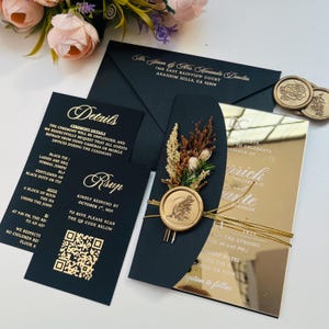 K&ouml;nnte beinhalten: Schwarze und goldene Hochzeitseinladung mit einem goldenen Siegel aus Wachs und einem Strau&szlig; getrockneter Blumen. Die Einladung hat eine spiegelnde Oberfl&auml;che und den Text "DC" und "Zur Feier der Hochzeit von" in goldenen Buchstaben. Die Namen "William II" und "Marite Johnson" sind ebenfalls in goldenen Buchstaben.