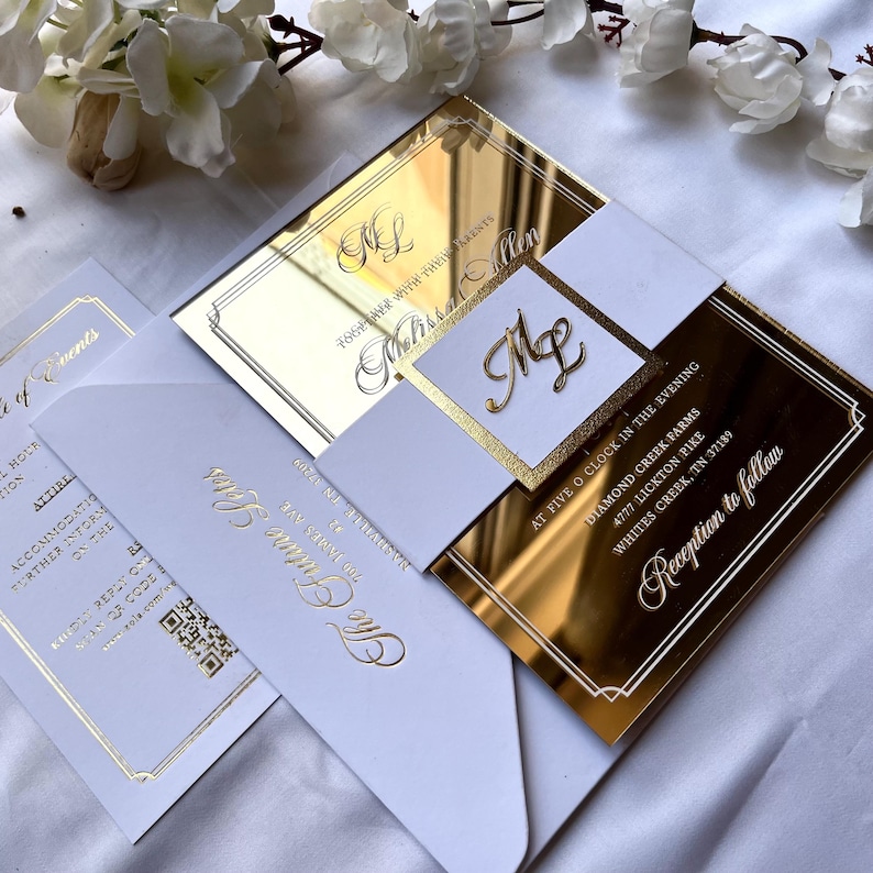 K&ouml;nnte beinhalten: Goldene Spiegel-Hochzeitseinladung mit wei&szlig;en Akzenten. Die Einladung zeigt die Namen des Paares und das Hochzeitsdatum sowie die Uhrzeit und den Ort der Zeremonie und des Empfangs. Die Einladung ist mit einem goldenen Rahmen und einem goldenen Monogramm verziert.