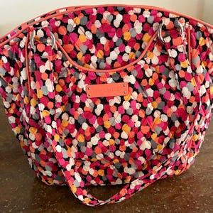 May include: A quilted tote bag with a colorful pattern of pink, orange, gray, and white dots on a black background. The bag has a coral-colored trim, handles, and a small tag that reads "VERA BRADLEY".