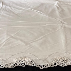 May include: A rectangular white tablecloth featuring a smooth central fabric panel and an intricate, scalloped lace border along the edges. The lace trim displays a detailed openwork pattern, providing a decorative finish to the plain white textile.