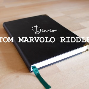 Puede incluir: Un diario de cuero negro encuadernado con detalles dorados y un marcapáginas de cinta verde. La portada dice "Diario" y "TOM MARVOLO RIDDLE".