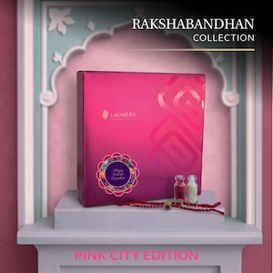 May include: A bright pink square gift box labeled RAKSHABANDHAN COLLECTION and LAUMIERE, featuring a circular floral emblem with the text Happy Raksha Bandhan. Two small glass vials and a red thread bracelet with a central stone are placed in front.