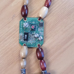 May include: A green circuit board with brown and cream wooden beads and two brown leather straps with metal accents. The circuit board has the number "3002" printed on it.
