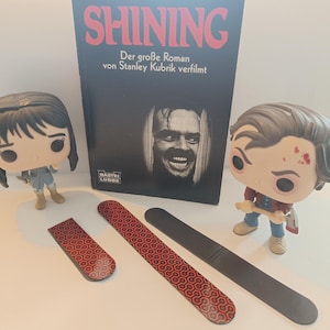Könnte beinhalten: Eine Sammlung von Artikeln zum Thema "Shining" von Stephen King. Das Bild zeigt ein Buch mit dem Titel "Shining", zwei Funko Pop Figuren und drei rechteckige Magnete. Die Magnete haben ein rotes und schwarzes geometrisches Muster. Das Buch zeigt Jack Nicholson.