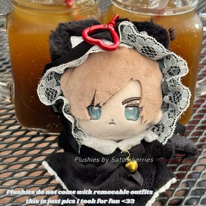 May include: A plush doll with light brown hair and blue eyes, wearing a black dress with white lace trim and a small gold bell. A red heart-shaped clip is attached to the top. Text on the image reads: Plushies by Saturnberries.
