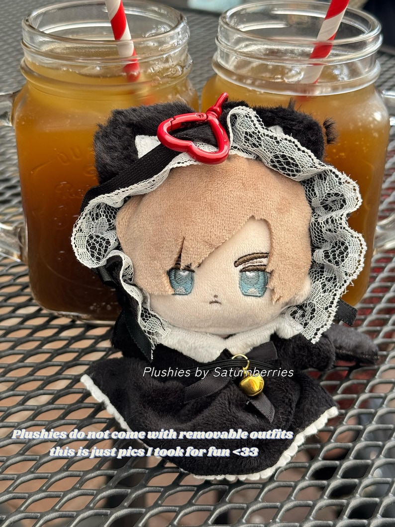 May include: A plush doll with light brown hair and blue eyes, wearing a black dress with white lace trim and a small gold bell. A red heart-shaped clip is attached to the top. Text on the image reads: Plushies by Saturnberries.
