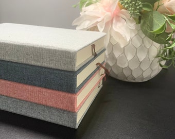 Peut inclure: Une pile de quatre journaux à couverture rigide avec des couvertures en tissu gris, bleu marine et corail. Chaque journal a des bords de pages crème et un signet en ruban marron. Un arrangement floral décoratif est en arrière-plan.