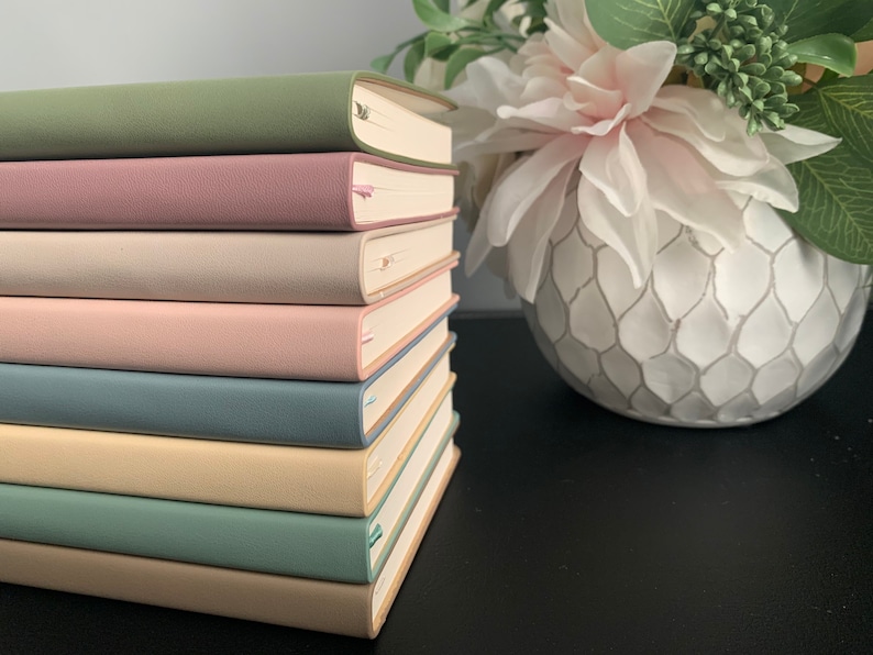 Peut inclure: Une pile de huit cahiers avec des couvertures de diff&eacute;rentes couleurs. Les couvertures sont vertes, roses, beiges, roses p&acirc;les, bleues, beiges, vertes et beiges. Les cahiers sont empil&eacute;s sur une surface noire.
