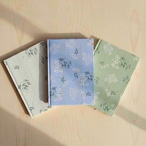 May include: Three notebooks with floral patterns. One is light blue with vertical stripes and white flowers, another is light green with white flowers, and the third is a light grey with white flowers. The notebooks are arranged on a light wooden surface.