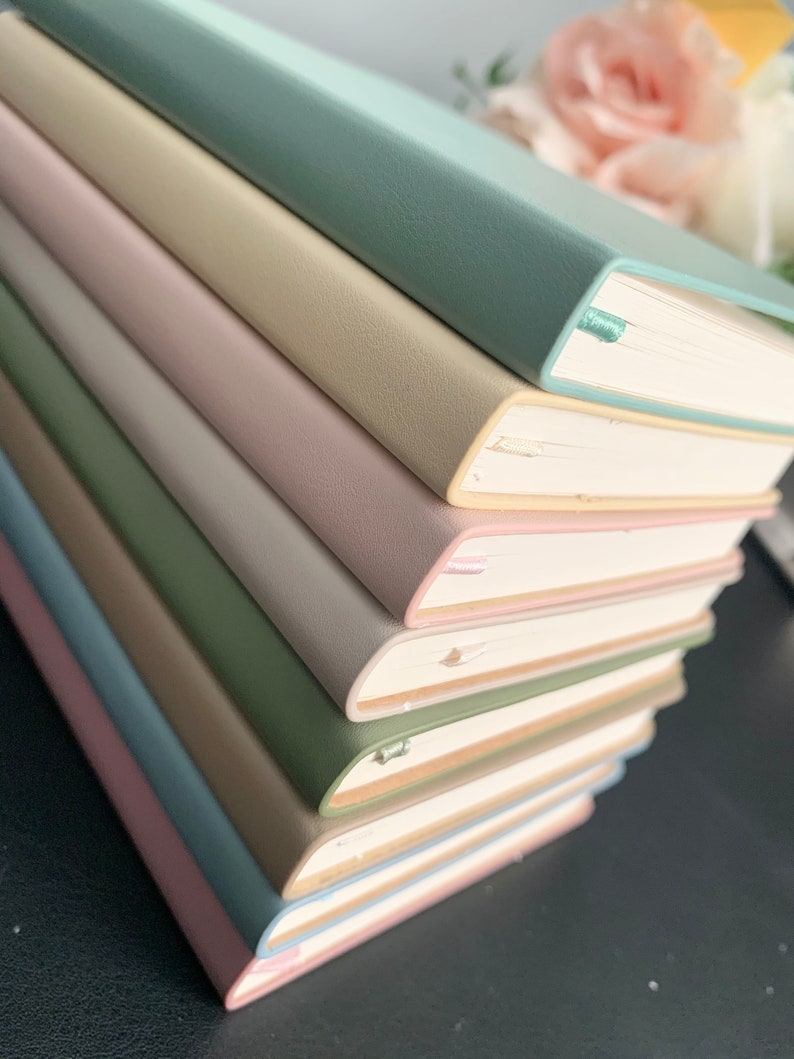 Peut inclure: Une pile de huit cahiers avec des couvertures de diff&eacute;rentes couleurs. Les couvertures sont bleu clair, vert clair, beige, rose, blanc, vert et bleu fonc&eacute;. Les cahiers sont empil&eacute;s sur une surface noire.