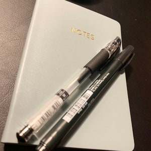 Peut inclure: Un carnet bleu clair avec le mot "NOTES" imprim&eacute; en lettres dor&eacute;es. Deux stylos sont pos&eacute;s sur le carnet, un noir et un transparent.