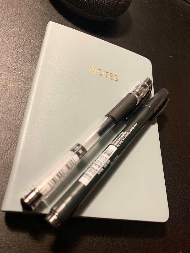 Peut inclure: Un carnet bleu clair avec le mot "NOTES" imprim&eacute; en lettres dor&eacute;es. Deux stylos sont pos&eacute;s sur le carnet, un noir et un transparent.