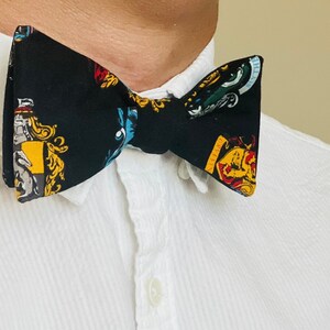 May include: A black bow tie featuring colorful crests and emblems. The bow tie is worn against a white collared shirt. The bow tie's design includes yellow, red, blue, and green accents, creating a striking contrast against the black fabric.