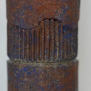 May include: A cylindrical ceramic vase with a textured surface. The vase is brown with blue accents and has a rough, rustic look.