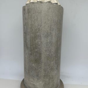 May include: A cylindrical, gray concrete garden ornament. The base and top are a darker shade of gray. The top has a decorative, ruffled edge in a lighter color, and is topped with a small, rounded element.