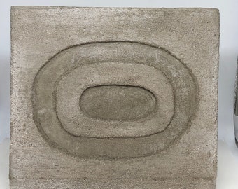 May include: A square, gray concrete tile with a raised, concentric oval design.