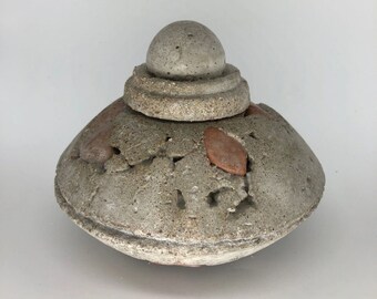 Può includere: Un vaso decorativo in cemento grigio con un coperchio. Il vaso ha un coperchio rotondo a forma di cupola con una superficie testurizzata. Il vaso ha una superficie testurizzata con un motivo di piccole forme irregolari. Il vaso ha un colore marrone chiaro.
