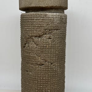 Puede incluir: Una escultura cilíndrica de hormigón texturizado con una parte superior ancha y plana. El cuerpo de la escultura tiene un patrón repetitivo de pequeños cuadrados. El color es un marrón apagado y terroso.