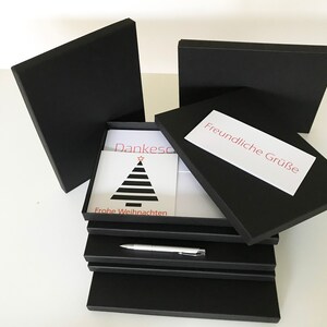 Puede incluir: Una pila de cajas de regalo negras con una tarjeta blanca en el interior. La tarjeta tiene un dise&ntilde;o de &aacute;rbol de Navidad rojo y negro y el texto "Dankesch&ouml;n" y "Frohe Weihnachten". La caja superior tiene una etiqueta blanca con el texto "Freundliche Gr&uuml;&szlig;e".
