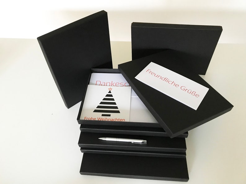 Puede incluir: Una pila de cajas de regalo negras con una tarjeta blanca en el interior. La tarjeta tiene un dise&ntilde;o de &aacute;rbol de Navidad rojo y negro y el texto "Dankesch&ouml;n" y "Frohe Weihnachten". La caja superior tiene una etiqueta blanca con el texto "Freundliche Gr&uuml;&szlig;e".