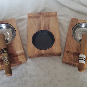 May include: Three wooden cigar rests with metal ashtrays. Each rest has a slot for a cigar and a round ashtray. The rests are made of light brown wood with a natural finish. The ashtrays are made of polished metal. The cigars are not included.