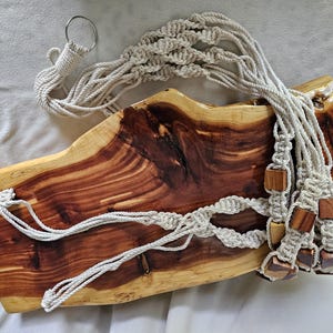 May include: A handcrafted macrame plant hanger with a natural wood base. The beige cotton rope is knotted in a decorative pattern, with wooden beads at the bottom. The wood base has a rich, reddish-brown grain.
