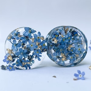 Puede incluir: Par de medidores de oreja de resina transparente con flores azules de nomeolvides y escamas doradas incrustadas en el interior.