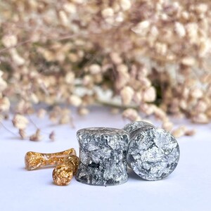 May include: Two gray and silver ear gauges with a silver flake design. There is a smaller gold ear gauge with gold flakes in the background.