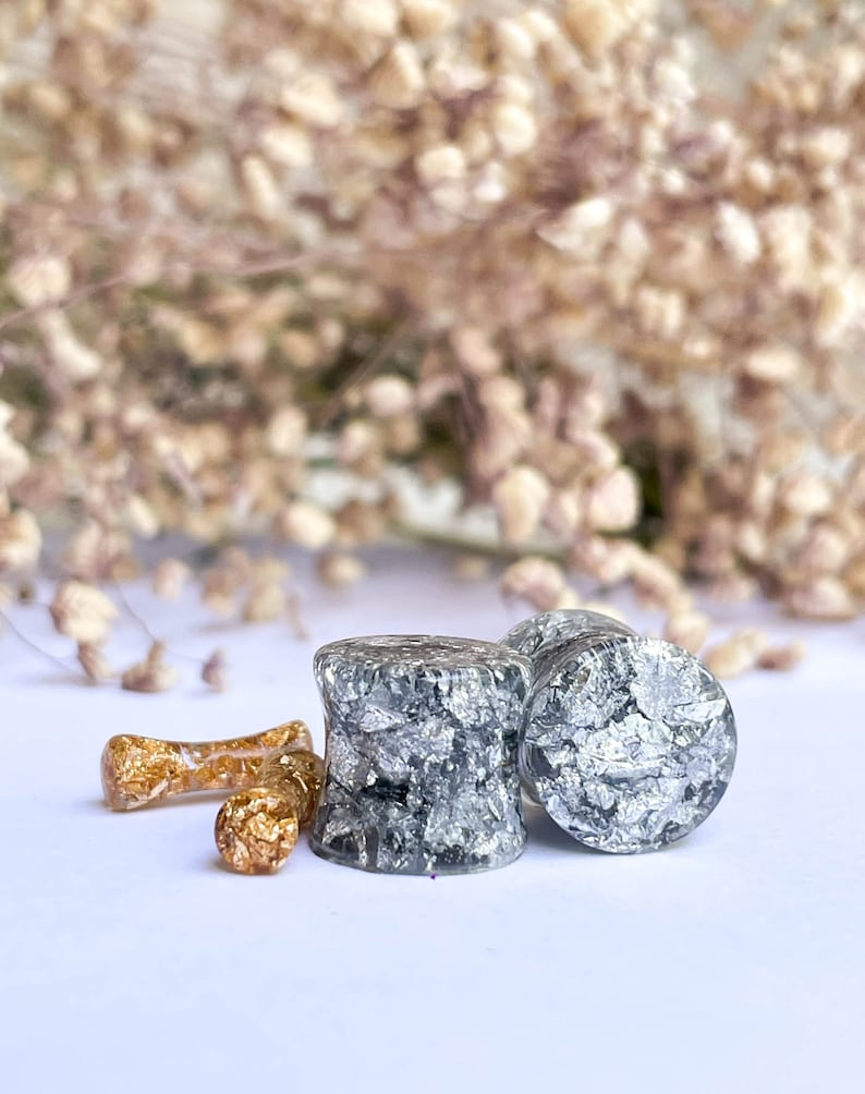 May include: Two gray and silver ear gauges with a silver flake design. There is a smaller gold ear gauge with gold flakes in the background.