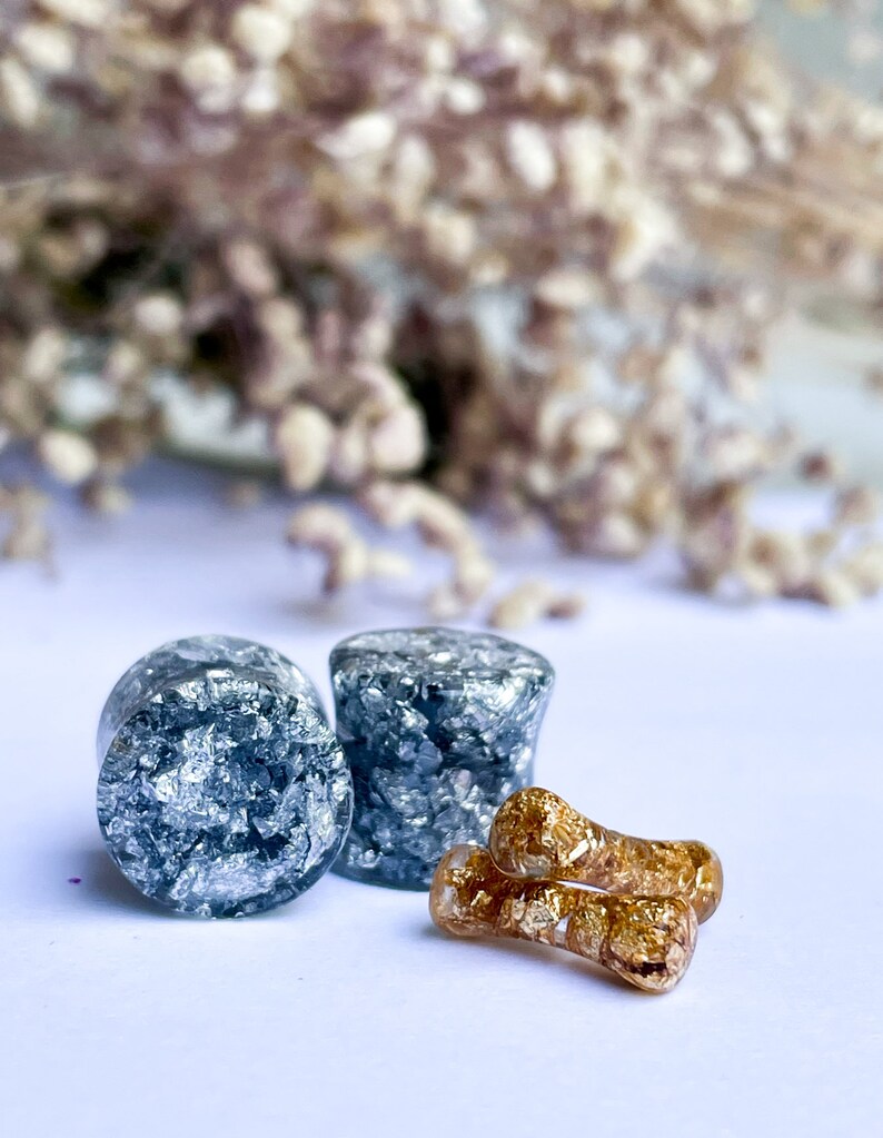 May include: Two round silver and black ear gauges with a pair of gold and black ear gauges.