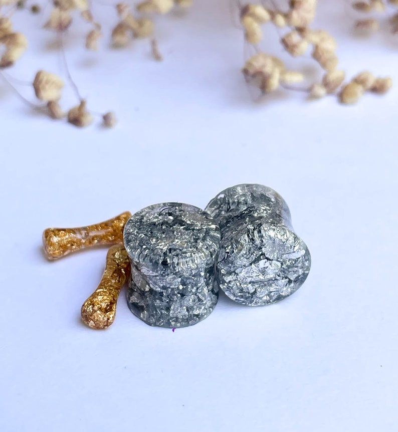 May include: Two silver and black ear gauges with a speckled pattern. The gauges are made of clear resin with silver flakes embedded in it.