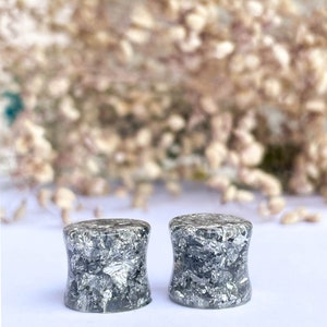 May include: A pair of black and silver ear gauges with a speckled pattern. The gauges are made of resin and have a cylindrical shape.