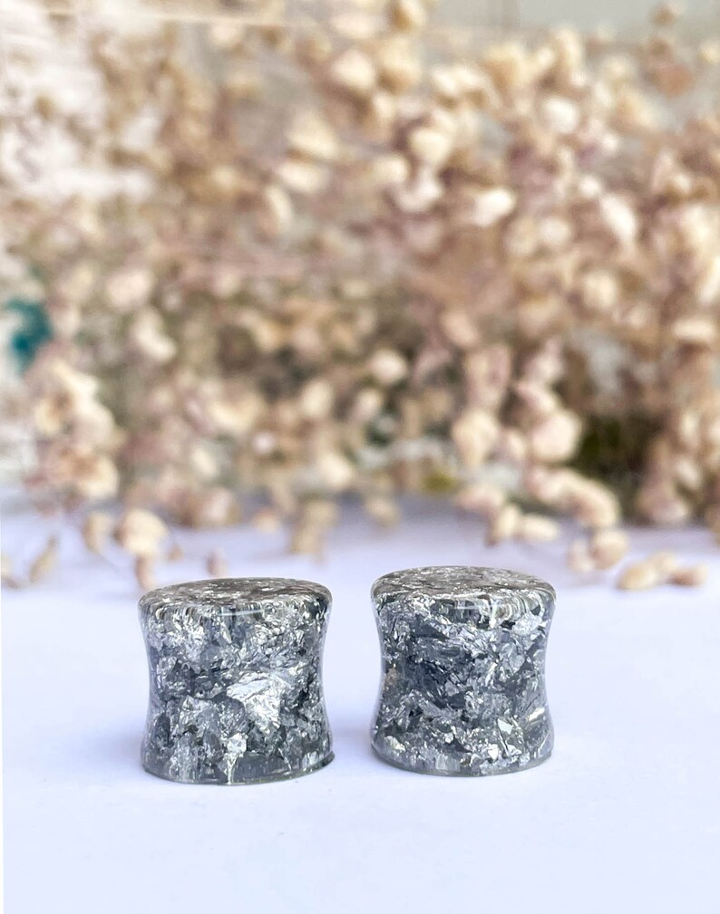 May include: A pair of black and silver ear gauges with a speckled pattern. The gauges are made of resin and have a cylindrical shape.