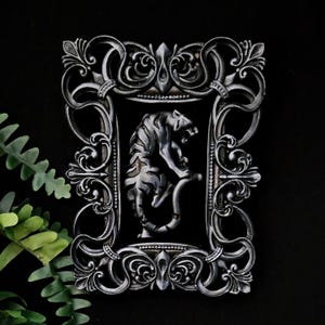 May include: A rectangular wall plaque featuring a silver-toned tiger in a crouching position, centered within an ornate, dark silver frame with intricate scrollwork and filigree details. The tiger is depicted in profile against a black background.