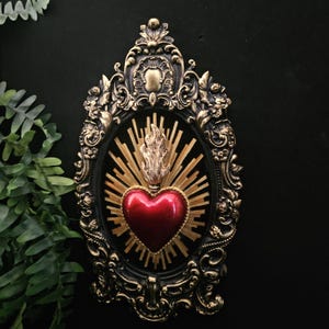 Puede incluir: Adorno de pared enmarcado y ornamentado. Un corazón rojo con una llama dorada y un diseño de rayos de sol se encuentra sobre un óvalo negro, enmarcado por un borde negro detallado con detalles dorados. Decoración gótica.