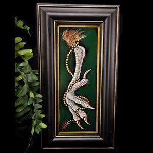 May include: Framed artwork featuring a detailed illustration of a bird's foot with sharp talons. The artwork is set against a dark green background with gold accents, and is enclosed in a black frame. The bird's foot is painted in shades of gray, pink, and white.