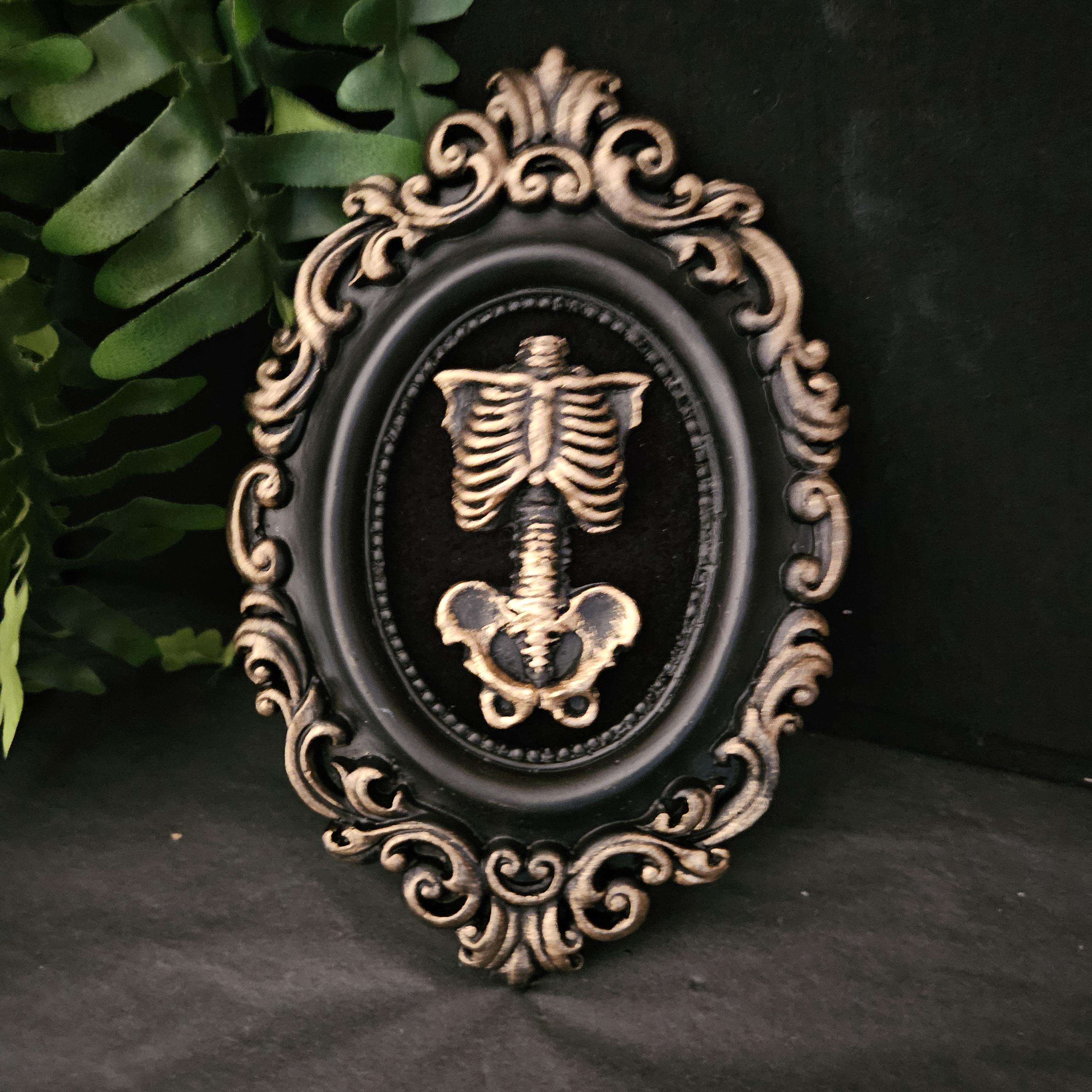 Burnished gold hand cast skeleton torso, rib cage, in a black and gold oval ornate frame, mounted on black velvet. Gothic art.