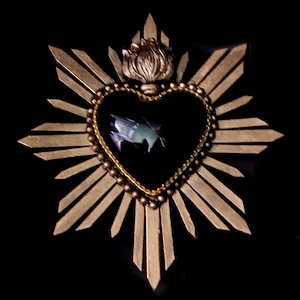 May include: A decorative wall ornament shaped like a heart with a black center, surrounded by a gold-toned sunburst frame. A sculpted gold flame sits atop the heart, which is bordered by a beaded gold trim and a thin gold chain detail.
