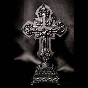 May include: A detailed, dark grey crucifix with an ornate design. The cross features a figure of Jesus and is mounted on a decorative base. The overall aesthetic is gothic and religious.